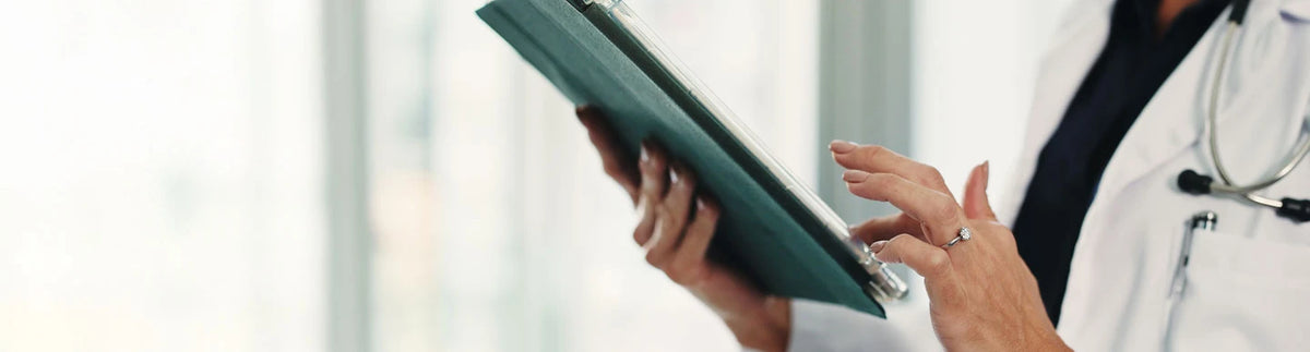 A close-up lifestyle shot of a doctor in a white coat holding a green clipboard, with a stethoscope around the neck and hands visible against a bright, blurred office background.
